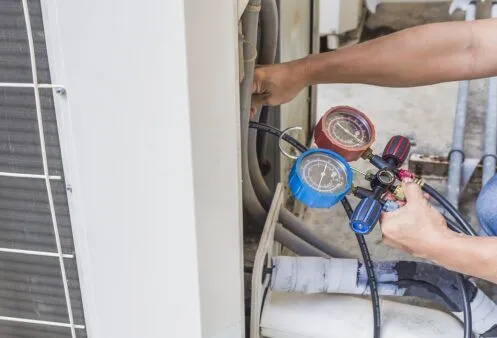 A technician is using a set of HVAC manifold gauges with blue and red dials to check the refrigerant levels on an outdoor air conditioning unit. The technician is adjusting the connected hoses while monitoring the pressure readings.