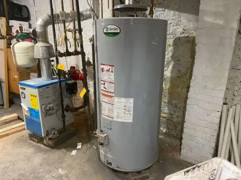 A water heater tank labeled "Smith" is installed in a basement next to a blue boiler unit, with various pipes and ventilation ducts connected. The surroundings include exposed brick walls and concrete flooring.