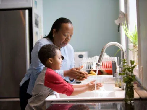 An adult helps a young child wash their hands at a kitchen sink, with water running from a modern faucet. Sunlight streams in from a window, and kitchen items such as dishes and plants are visible on the countertop.