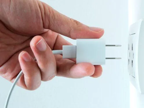 A hand is plugging a charger into a wall outlet, preparing to connect the device to power. The charger is a standard two-prong plug with a white casing.