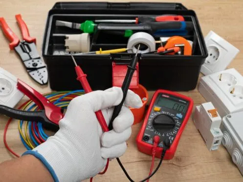 A person wearing a protective glove is holding multimeter probes in front of a toolbox filled with electrical tools, including wire cutters, electrical tape, and cables. Various electrical outlets and components are arranged around the workspace.