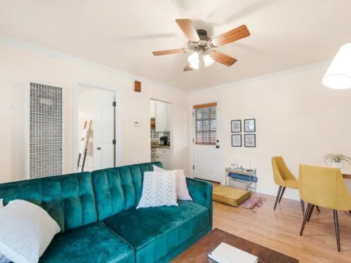 A bright living room features a teal velvet sofa, a ceiling fan with wooden blades, and a small dining area with yellow chairs. The room has hardwood floors, white walls, and access to a kitchen through an open door.