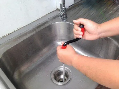 A person is using a hand crank drain auger to unclog a kitchen sink by inserting the tool into the drain. The stainless steel sink and faucet are visible, with a metal drain strainer in place.