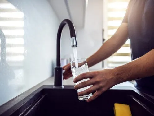 A person is filling a glass of water from a modern kitchen faucet, with water streaming into the glass. The setting includes a dark sink and bright lighting coming from a nearby window.