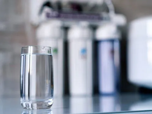 A clear glass of water sits on a countertop in front of a blurred background of a water filtration system. The setup suggests purified drinking water from a home filtration unit.