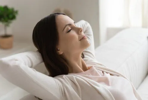 A woman relaxes on a couch with her hands behind her head and eyes closed, enjoying a moment of peace and comfort. Soft lighting and a neutral background contribute to the tranquil atmosphere.