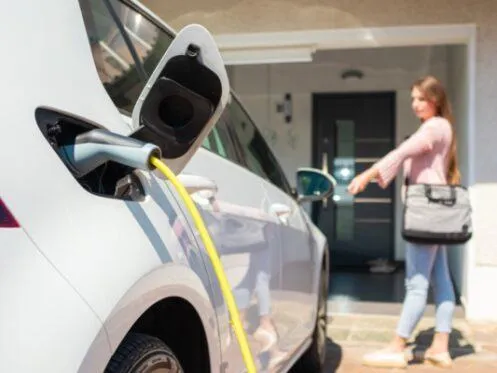 An electric vehicle is charging in a driveway, with a yellow charging cable plugged into the car. In the background, a woman with a bag is walking toward the house.