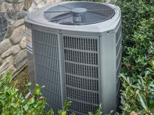 An outdoor central air conditioning unit is placed next to a stone wall, surrounded by green bushes. The unit's fan and vented sides are visible, indicating it is part of a home HVAC system.