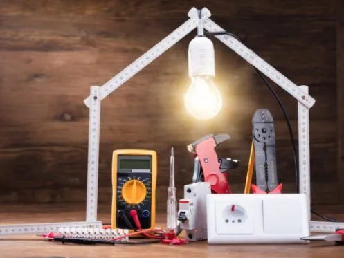 A collection of electrical tools, including a multimeter, wire cutters, and a power outlet, is arranged under a glowing lightbulb framed by a folding ruler in the shape of a house. The scene symbolizes electrical work and home wiring.