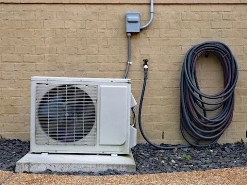 An outdoor HVAC unit is installed against a brick wall, with coiled hoses mounted next to it and electrical wiring running into a power box above. The unit sits on a concrete slab, surrounded by mulch and a gravel pathway.