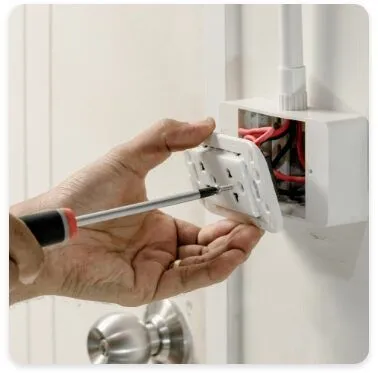 A person is using a screwdriver to install or remove the faceplate of an electrical outlet, with exposed wiring visible inside the electrical box. The surrounding area includes a door and a metal door handle.