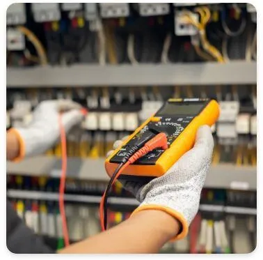 An electrician wearing protective gloves is using a digital multimeter to test electrical circuits in a control panel filled with wires and breakers. The multimeter probes are connected to measure voltage or continuity.