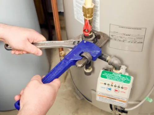 A person is using two wrenches to adjust a valve on the top of a water heater. The valve and surrounding components are part of the water heater's piping system.