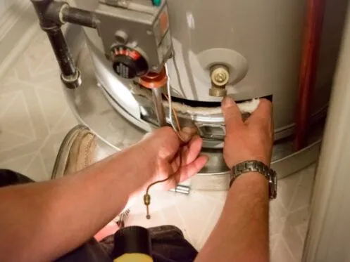 A person is repairing the bottom section of a water heater, using a tool to adjust or tighten a component. The water heater shows signs of damage or wear near the bottom, where the repair is taking place.