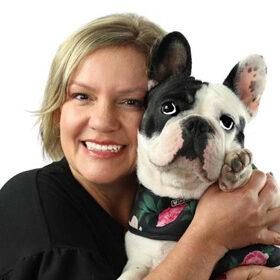 A smiling woman holds a French Bulldog close, both facing the camera. The dog wears a floral-patterned harness, and the woman has short blonde hair.